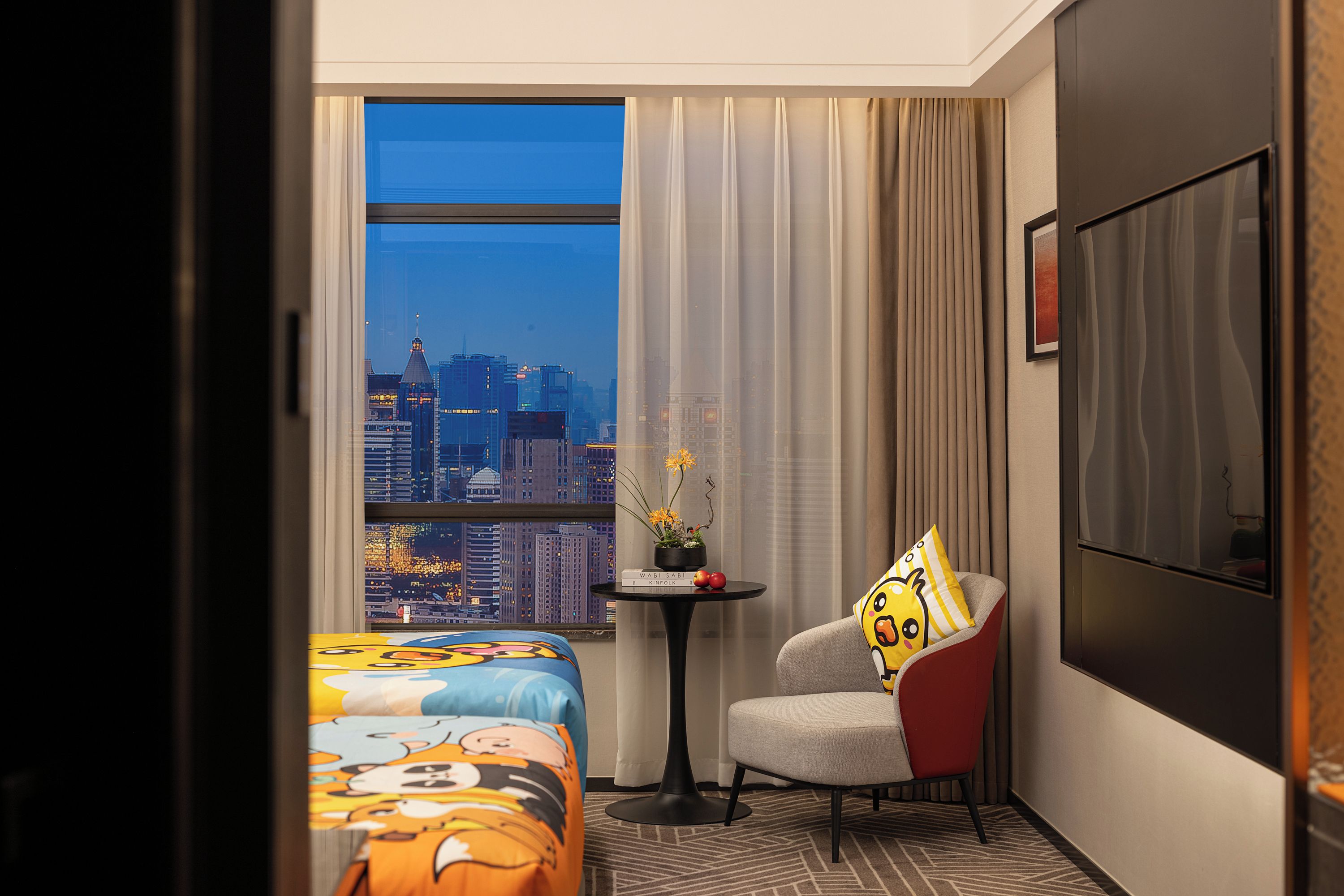 Ein Hotelzimmer mit einem Bett, einem Sessel, einem kleinen Tisch mit Pflanze und Blick auf die Stadt durch das Fenster.