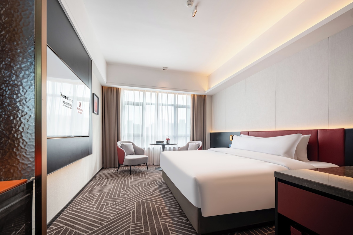 Une chambre d’hôtel avec un lit, un bureau, deux chaises, une fenêtre avec des rideaux et une moquette à motifs.