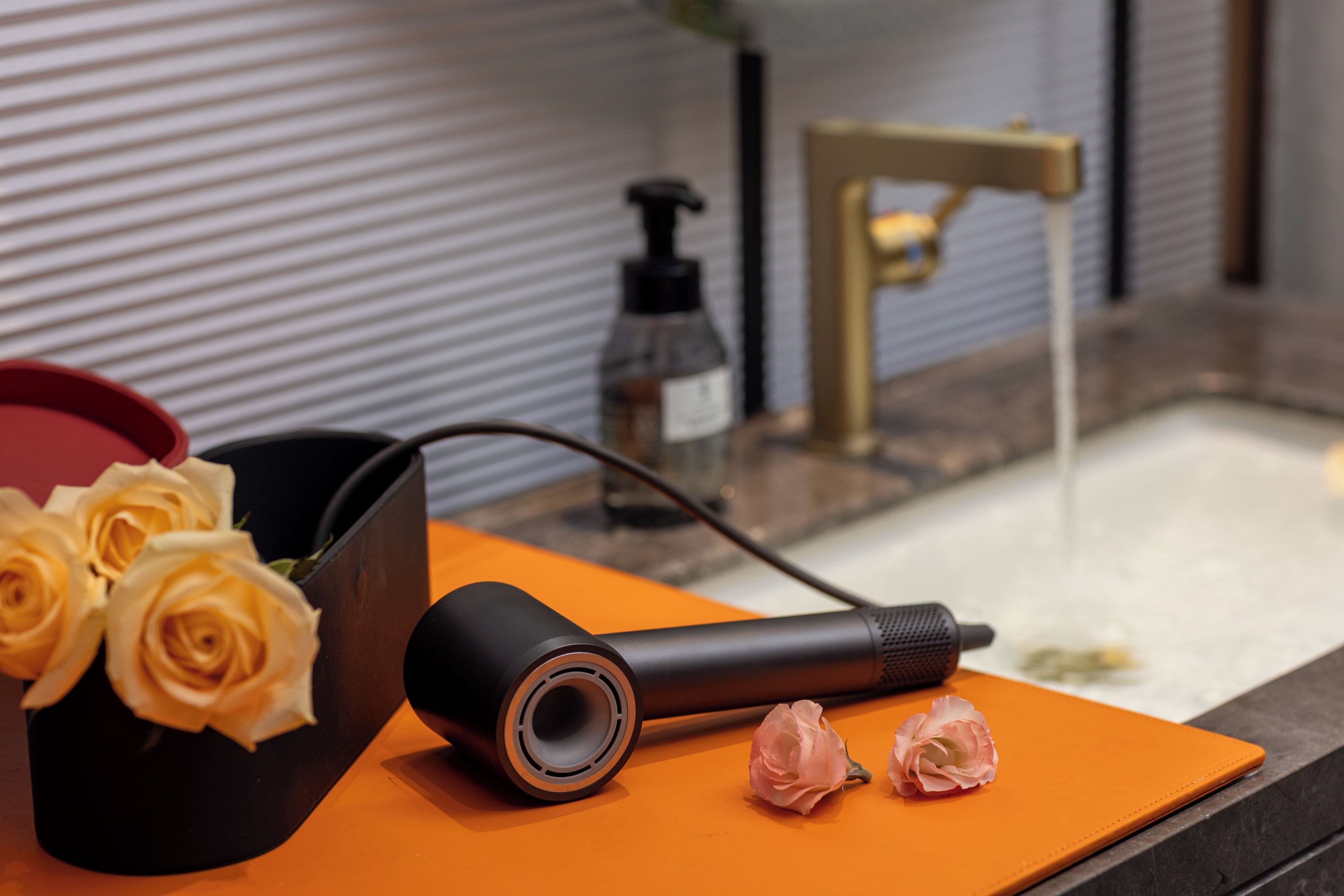 Un baño con un secador negro, un recipiente rojo con rosas amarillas y un grifo dorado.