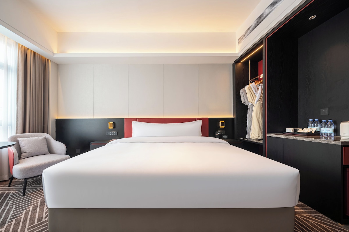 Une chambre d’hôtel avec un grand lit, une table de chevet, une chaise, une armoire et un bureau doté d’un miroir et d’articles de toilette.