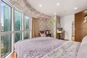 Ein Schlafzimmer mit großem Fenster, einem Bett mit gemustertem Überwurf, einem Schminktisch mit Spiegel und Blick auf die Stadtlandschaft.