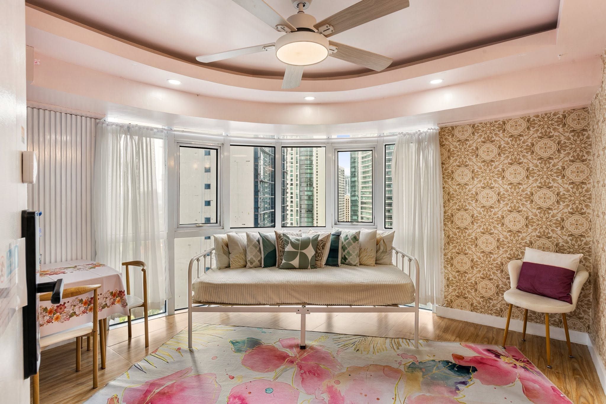 A room with a curved ceiling, a bed with multiple pillows, a chair, a table with a floral tablecloth, and a patterned rug.