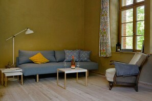 Ein Wohnzimmer mit einer Couch, einem kleinen Tisch, einer Lampe, einem Sessel und einem Fenster mit Vorhängen.