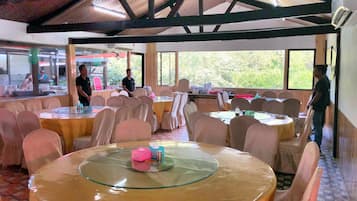 A restaurant with round tables and chairs, large windows, and a high ceiling with wooden beams.