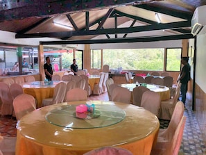 Un restaurante con mesas y sillas redondas, amplios ventanales y un techo alto con vigas de madera.