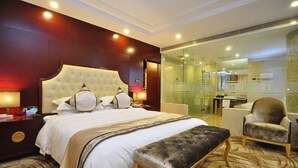 Een hotelkamer met een groot bed, een zithoek met een bankje, een stoel, een douchecabine met glazen wanden en een klein bureau met lamp.
