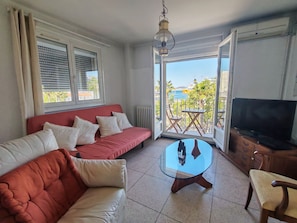 Ein Wohnzimmer mit einem roten Sofa, einem Glastisch, einem Fernseher und einem Balkon mit Meerblick.