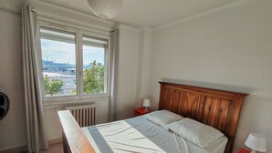Un dormitorio con una cama de madera, una ventana con vistas a un paisaje urbano, un radiador y una mesita de noche roja con una lámpara.