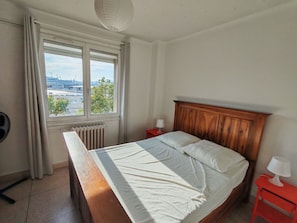 Ein Schlafzimmer mit einem hölzernen Bett, einem Fenster mit Blick auf eine Stadtansicht, einem Heizkörper und einem roten Nachttisch mit einer Lampe.