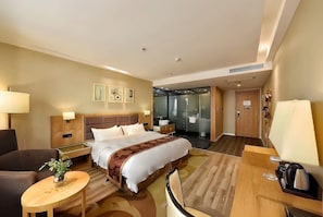 Ein Hotelzimmer mit Bett, Nachttischen, Schreibtisch, Sessel, Lampe, Pflanze und einer Duschkabine.