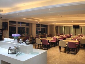 En restaurant i et hotel med runde borde og stole, et receptionsskrank samt et stort vindue.