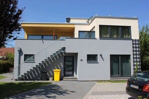 A modern two-story building with a grey facade, a visible staircase, and a car parked in the driveway. - Holiday apartment with 1 bedroom (Börgerende)