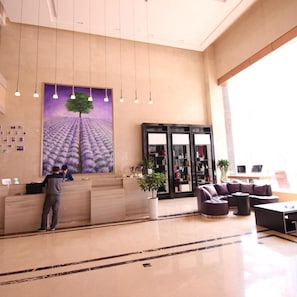 Un vestíbulo de hotel con mostrador de recepción, un cuadro grande de un campo de lavanda, un área de descanso con sofás y una estantería.