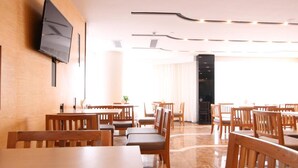 Ein Essbereich eines Hotels mit Holztischen und -stühlen, einem an der Wand befestigten Flachbildfernseher und einem modernen Deckenstil.
