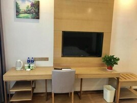 Una habitación con un escritorio de madera, un televisor montado en la pared, una silla, una pequeña planta en maceta y un cesto de basura.