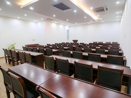 Un salón de conferencias con filas de mesas y sillas de madera, una pizarra blanca y una planta.