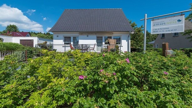 Une maison d’habitation avec un toit recouvert de tuiles, une véranda à l’avant, visible de la rue, avec une table et des chaises, et un panneau indiquant les coordonnées.