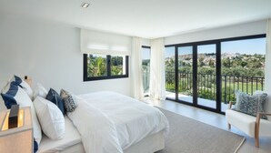 A modern bedroom with a large bed, a view of a green landscape, and a balcony with a railing. - Vrbo Property (Marbella)