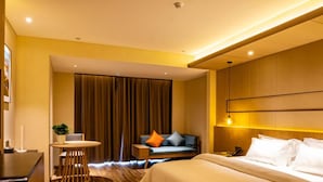 Ein Hotelzimmer mit einem großen Bett, einem Schreibtisch, einem Sessel, einem kleinen Tisch mit Pflanze und einer Couch mit Kissen.