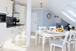 A modern kitchen with white cabinets, a dining area with a white table and chairs, and a clock on the wall. - Holiday apartment with 2 bedrooms (Zingst)