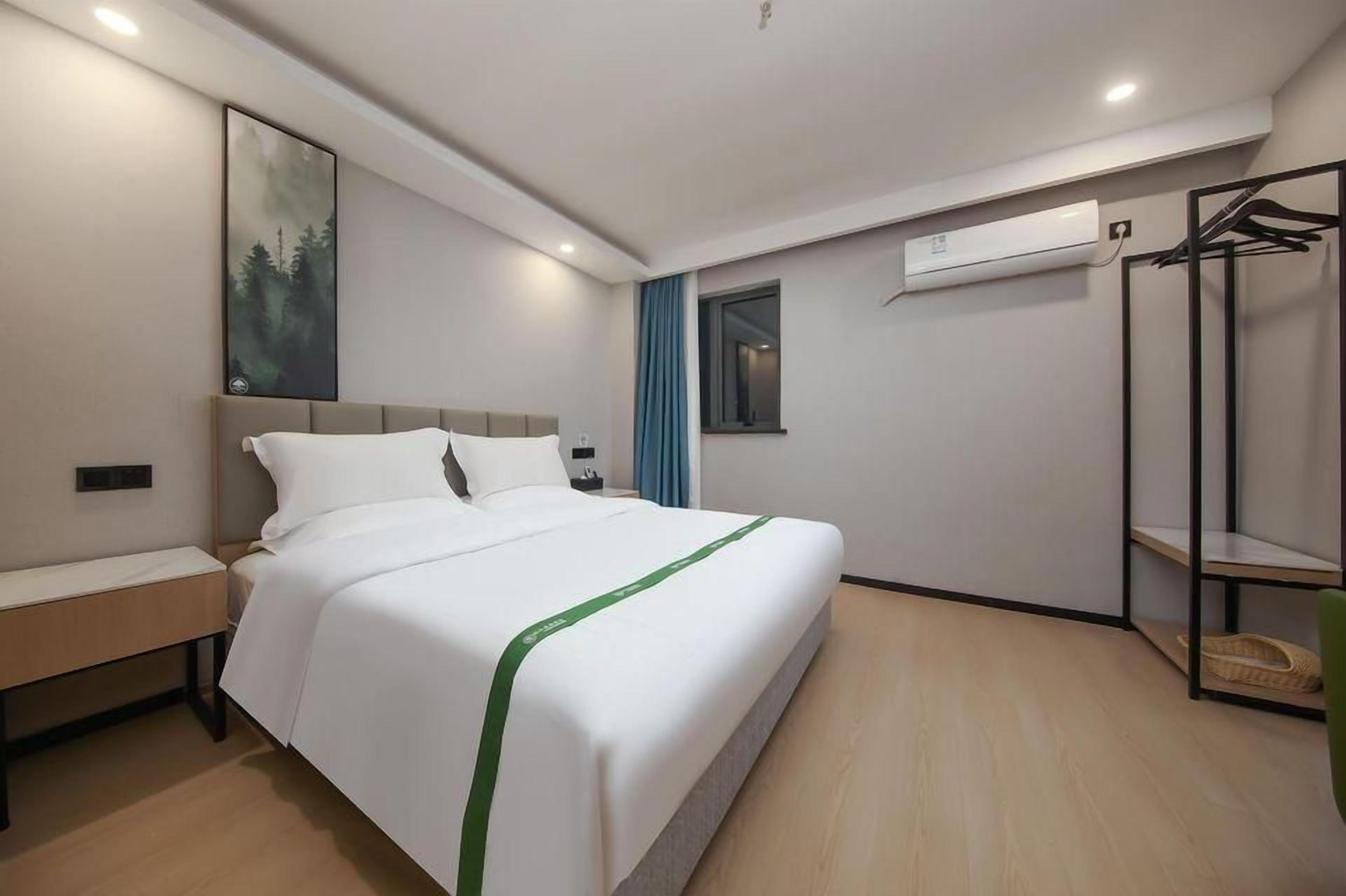 A modern hotel room with a large bed, a bench, a wall-mounted air conditioner, and a coat rack.