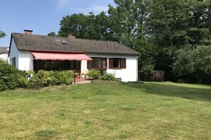 Ein eingeschossiges Haus mit einer roten Markise über dem Eingang, einem Schornstein und einem kleinen Gartenhaus im Hinterhof.