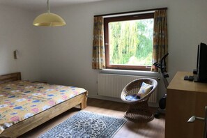 Ein Schlafzimmer mit einem Bett, einem Fenster mit Vorhängen, einem Korbstuhl, einer Heizung und einem Schreibtisch mit einem Computer.