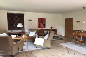 Ein Wohnzimmer mit Sofa, Ohrensessel, Couchtisch und einem Esstisch mit Stühlen. Es gibt ein Bücherregal, einen Kamin und einen großen Teppich auf dem Boden.