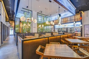 Interior moderno de restaurante con mesas y sillas de madera, lámparas colgantes y vistas a zonas verdes a través de amplios ventanales.
