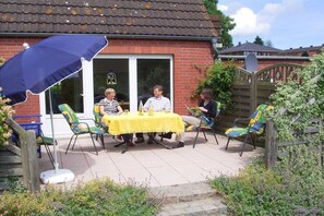 Eine Terrasse im Freien mit Tisch und Stühlen, einem blauen Sonnenschirm und einem Backsteinhaus im Hintergrund.