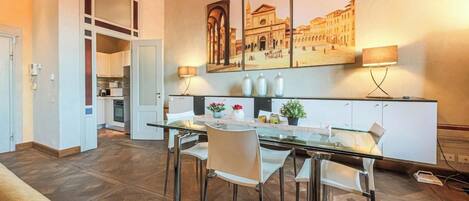 A modern kitchen with a dining area, featuring a glass table, white chairs, and a large cityscape painting on the wall.