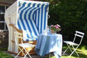 A wicker chair with a striped cushion, a small table with a blue tablecloth, and a white folding chair. - Holiday home in Oldsum with swimming pool (Oldsum)