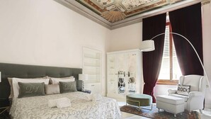 A bedroom with a large bed, a white armchair, a side table, a wardrobe, and a window with curtains. - Luxury Apartment in Central Florence (Florence)