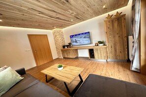 A modern living room with a wooden ceiling, a television mounted on the wall, a sofa, a coffee table with a plant, and a wooden cabinet. - Holiday apartment with 1 bedroom (St. Jakob)