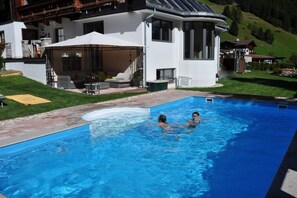 A swimming pool with two people in the water, surrounded by a white building with a covered patio and a view of a green hillside. - Holiday apartment with 1 bedroom (St. Jakob)