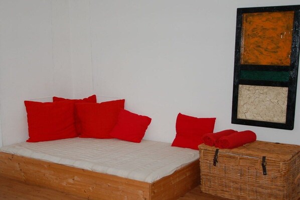 A single bed with a wooden frame and a white mattress, accompanied by several red pillows. A wicker basket with red towels is placed beside the bed. - Old Rectory Elmenhorst (Elmenhorst)