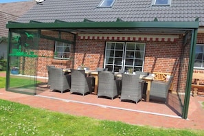A covered outdoor seating area with wicker chairs and a table, adjacent to a brick building with a window. - | Friesenhaus Deichperle (St. Peter-Ording)