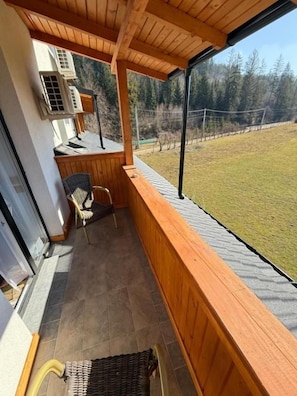 Un balcón de madera con vista a un área verde y árboles. Hay dos sillas de mimbre y una mesita.