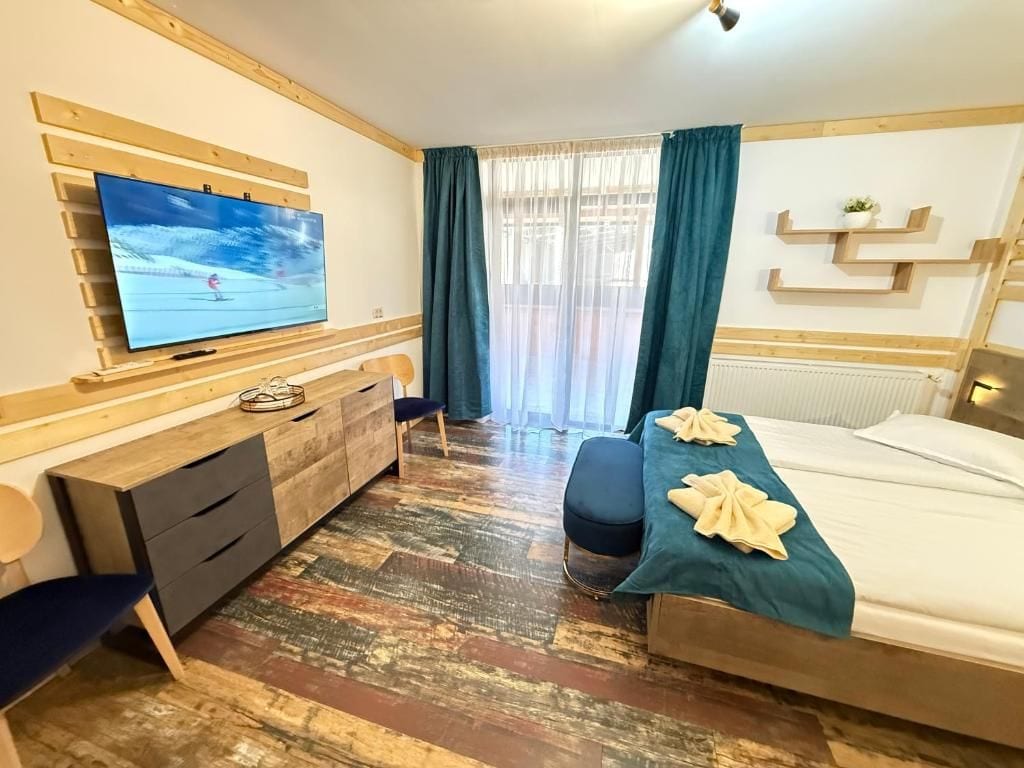A hotel room with a bed, a TV mounted on a wooden wall, a desk with drawers, a chair, and a window with curtains.
