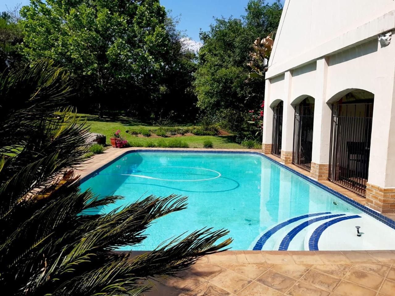 Una piscina con una fuente de agua azul cristalina, rodeada por un jardín ajardinado con palmeras y un edificio blanco con ventanas arqueadas.