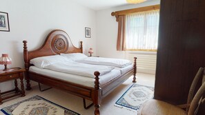 Ein ordentlich bezogenes Bett mit Holz-Kopfteil, ein Nachttisch mit Lampe und ein Fenster mit Vorhängen.