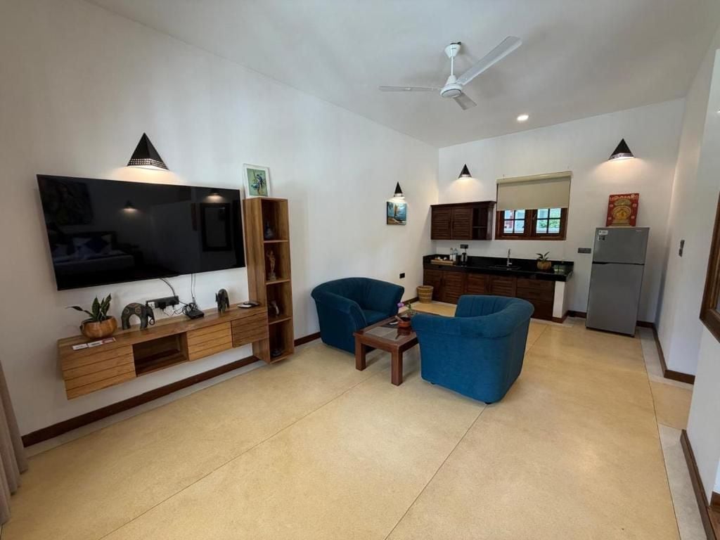 A living room with a flat-screen TV, a ceiling fan, and a kitchen area with cabinets and a refrigerator.