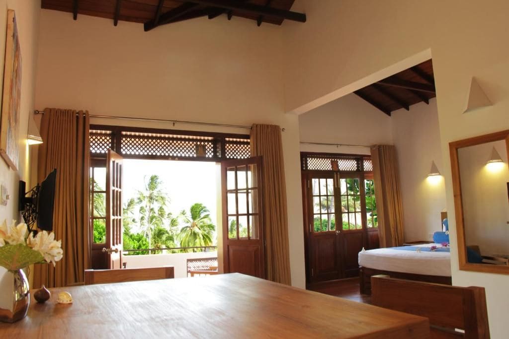 Una habitación con una mesa de madera, una cama y vistas a palmeras a través de puertas abiertas.
