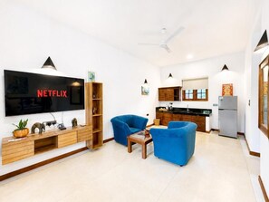 A modern living room with a flat-screen TV, blue armchairs, wooden entertainment unit, and a kitchen in the background.
