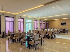 A spacious dining area with multiple tables and chairs, large windows with purple-tinted glass, and a flat-screen TV mounted on the wall. - Spree Resort at Century Wintersun (Bengaluru)