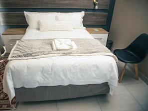 A neatly made bed with a white comforter and a grey blanket, a black chair, and a wooden nightstand with a lamp.