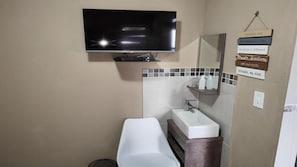 A bathroom with a flat-screen TV mounted on the wall, a white sink, and a mirror above it. There are toiletries on the sink's edge and a wall-mounted shelf. - Wilson Guest House (Groblersdal)