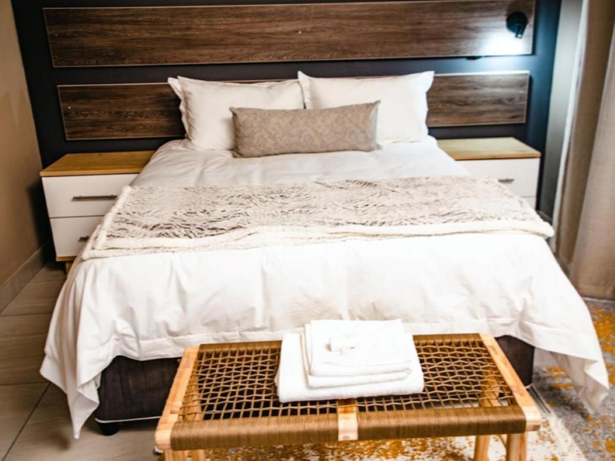 A neatly made bed with a white comforter and a decorative throw, a wooden nightstand with a drawer, and a woven stool with folded towels.