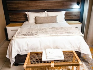 A neatly made bed with a white comforter and a decorative throw, a wooden nightstand with a drawer, and a woven stool with folded towels. - Wilson Guest House (Groblersdal)