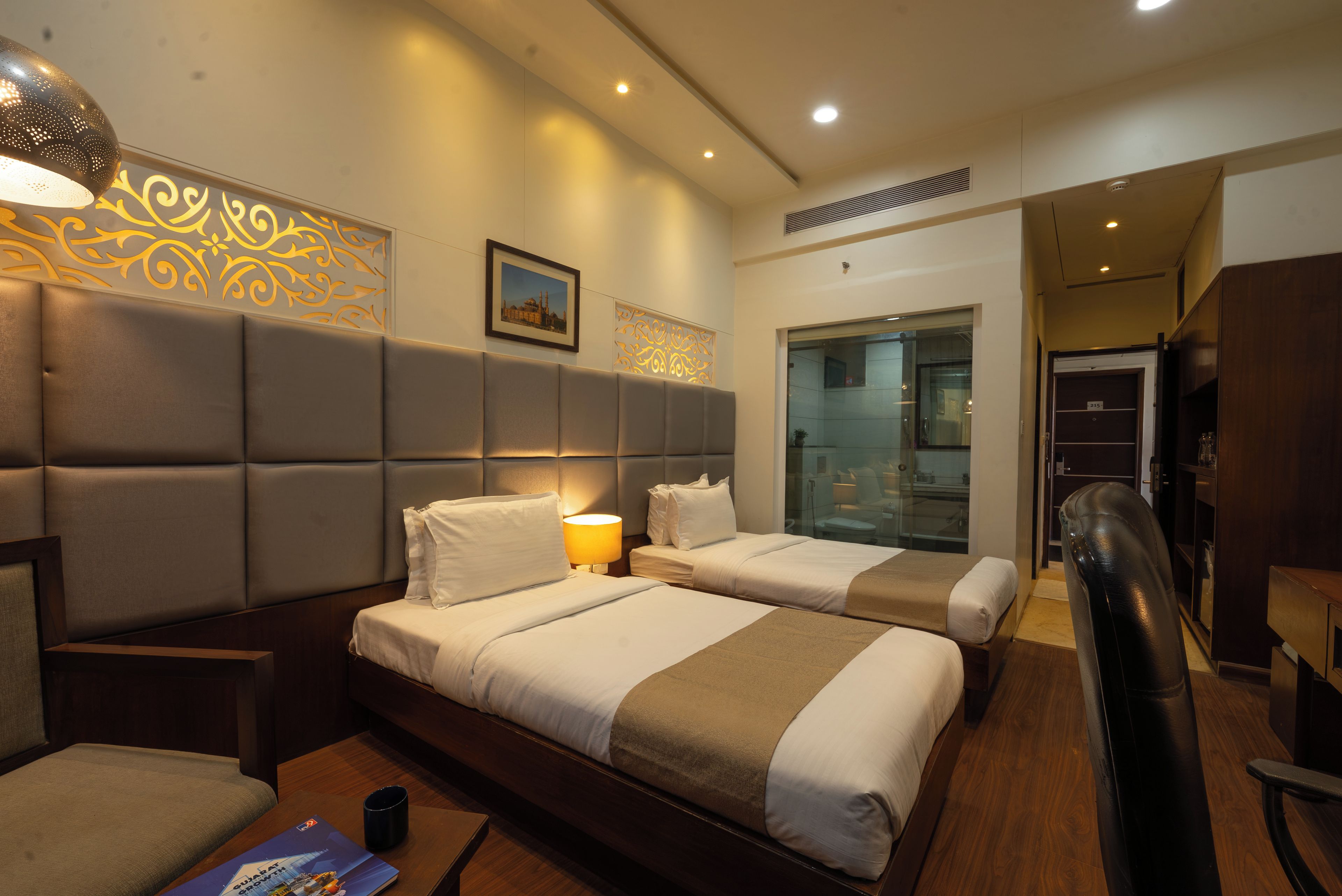 Habitación de hotel con dos camas, un sofá, un escritorio y un baño visible a través de una puerta de cristal.